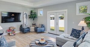 A modern living room with a flat-screen TV, two blue armchairs, a grey sofa, a coffee table with a bowl of popcorn, and a large window with a view of a green lawn. - 4BR Home With Fenced Yard & Grill (Panama City Beach)