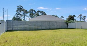 A fenced backyard with a grassy lawn, a building with a sloped roof, and trees in the background. - 4BR Home With Fenced Yard & Grill (Panama City Beach)