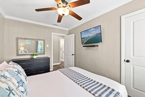 Ein Schlafzimmer mit Bett, Kommode, Spiegel, Deckenventilator, Fernseher und einer Tür.