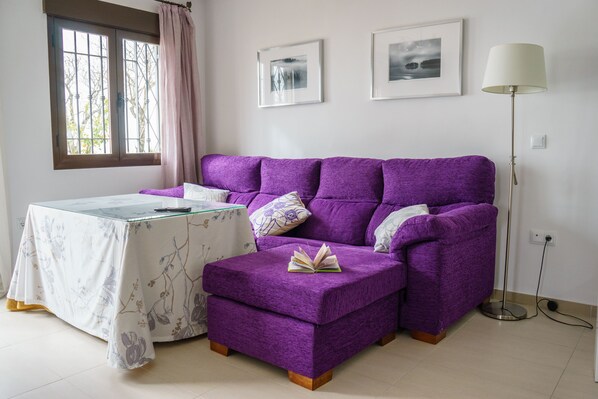 Ein Zimmer mit einem violetten Sofa, einem kleinen Tisch mit einer Glasplatte und zwei gerahmten Bildern an der Wand.