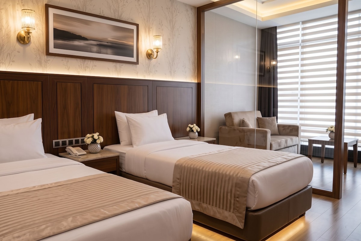 Habitación de hotel con dos camas, un cuadro sobre ellas, una mesita de noche con teléfono y jarrón, y una zona de estar con una silla y una mesa.