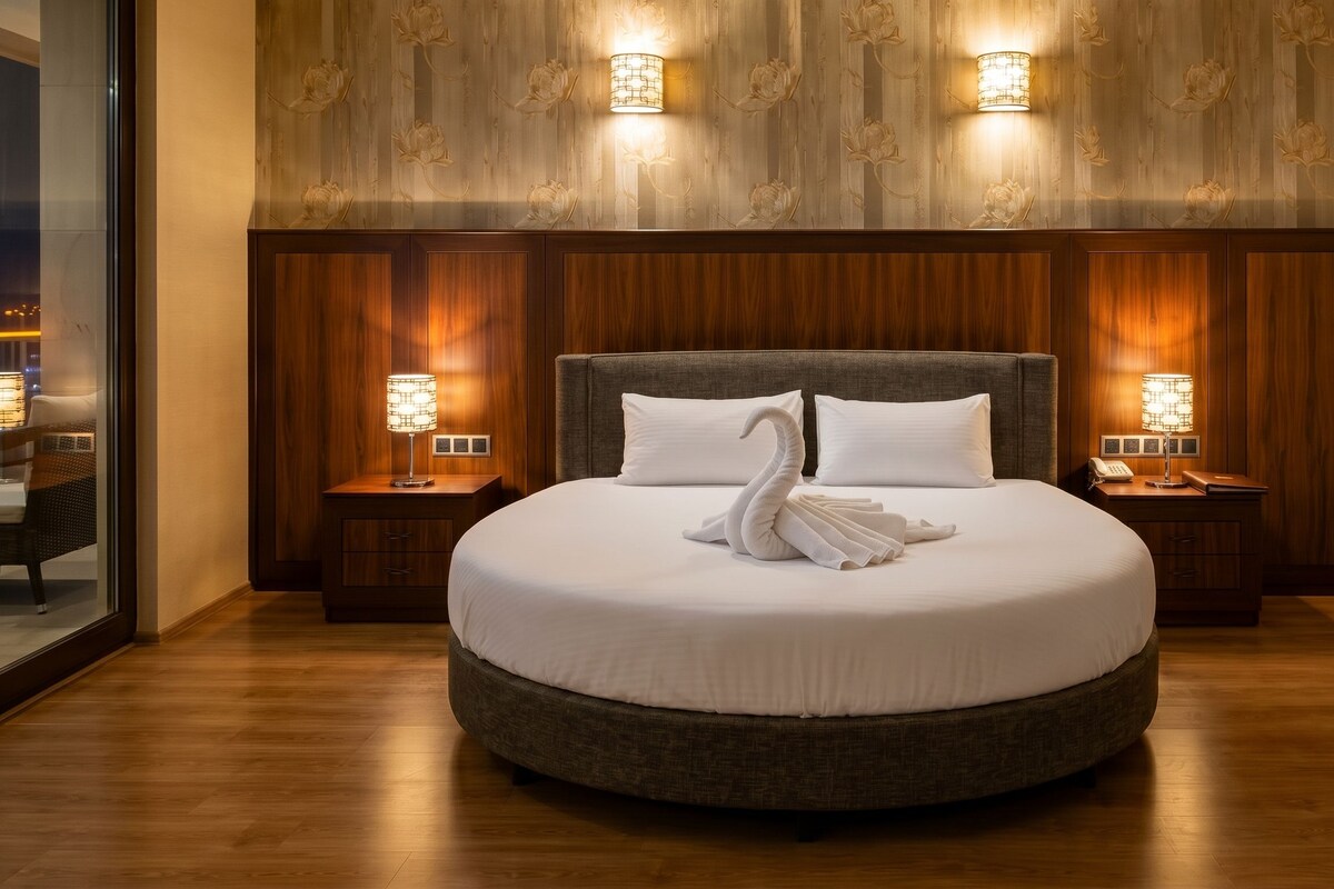 Una cama redonda con una escultura de cisne hecha con toallas, dos lámparas de noche y un panel de madera con estampado floral.
