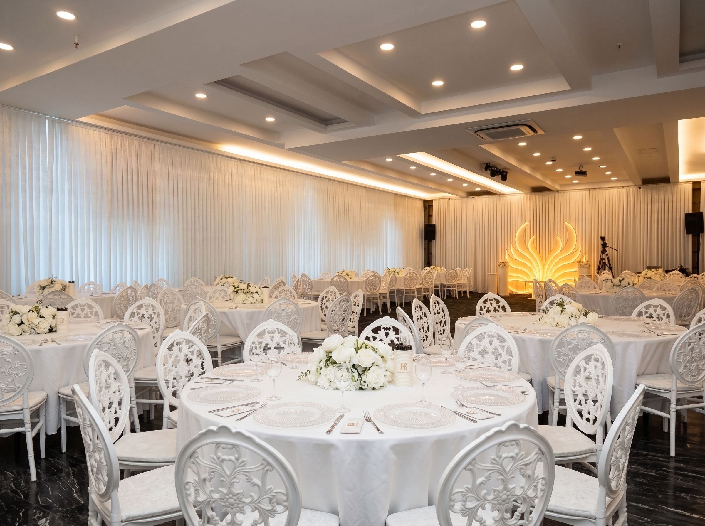 A banquet hall with round tables, white chairs, and floral centerpieces.
