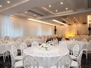 Un salón de banquetes con mesas redondas, sillas blancas y centros de mesa florales.