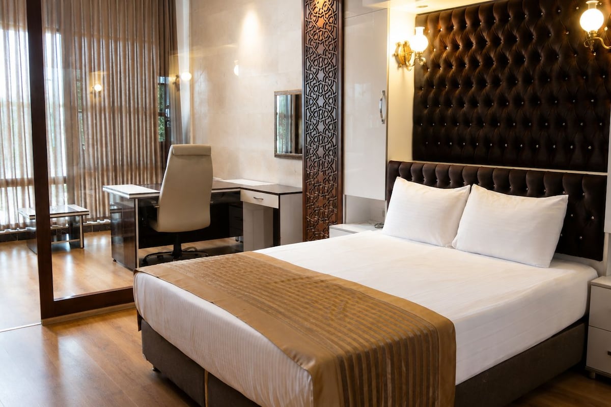 A hotel room with a large bed, a desk, and a chair. There is a window with curtains, a wall-mounted light fixture, and a decorative panel.
