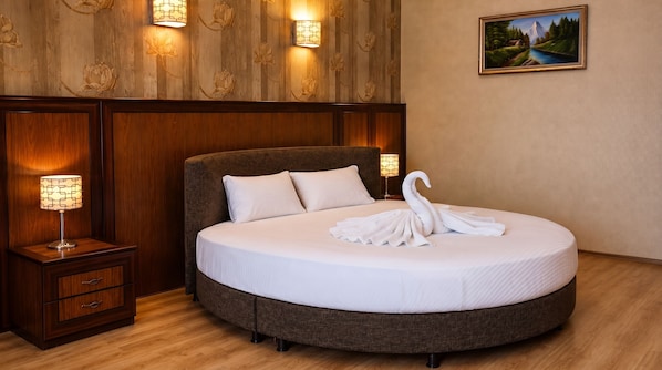 A hotel room with a round bed, two bedside lamps, a wooden headboard, a wall-mounted painting, and a wooden floor.