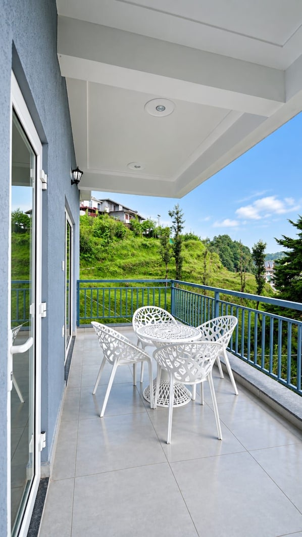 A balcony with white chairs and a table, a blue railing, and a view of a green hillside.