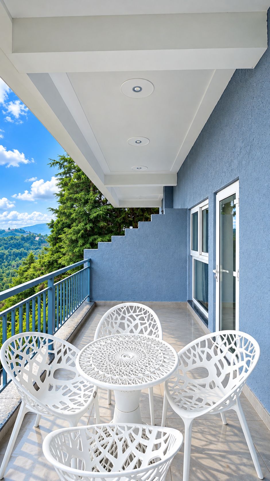 A balcony with white chairs and a table, a blue wall, and a view of a forested area.