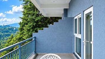 Un balcon avec des chaises blanches et une table, un mur bleu et une vue sur une zone boisée.