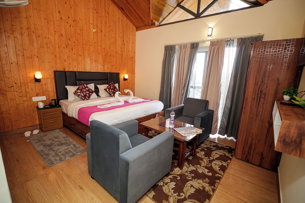A hotel room with a wooden interior, a bed with pillows, a bedside table with a bottle and glasses, a grey armchair, and a window with curtains.