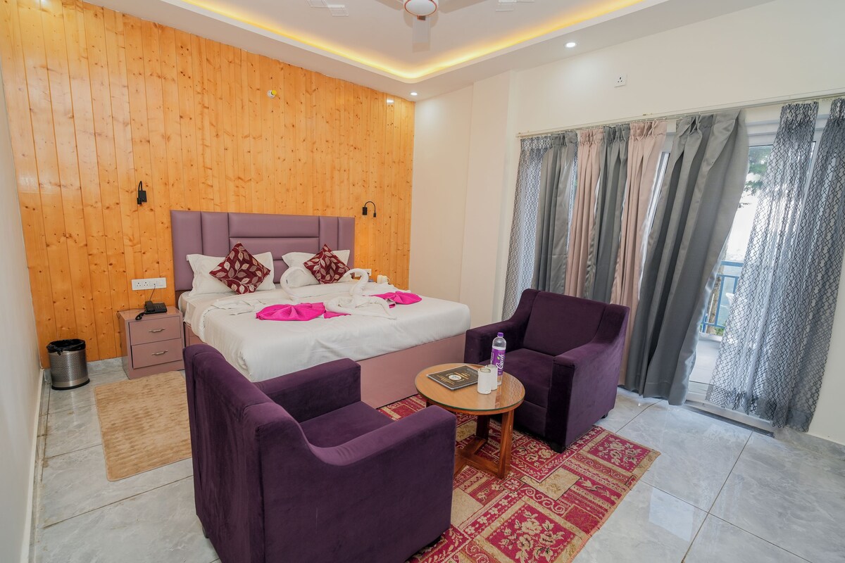 A modern hotel room with a wooden wall, a bed with white bedding and pink pillows, a purple armchair, a small table with a water bottle, and a view of the outdoors through a window with curtains.