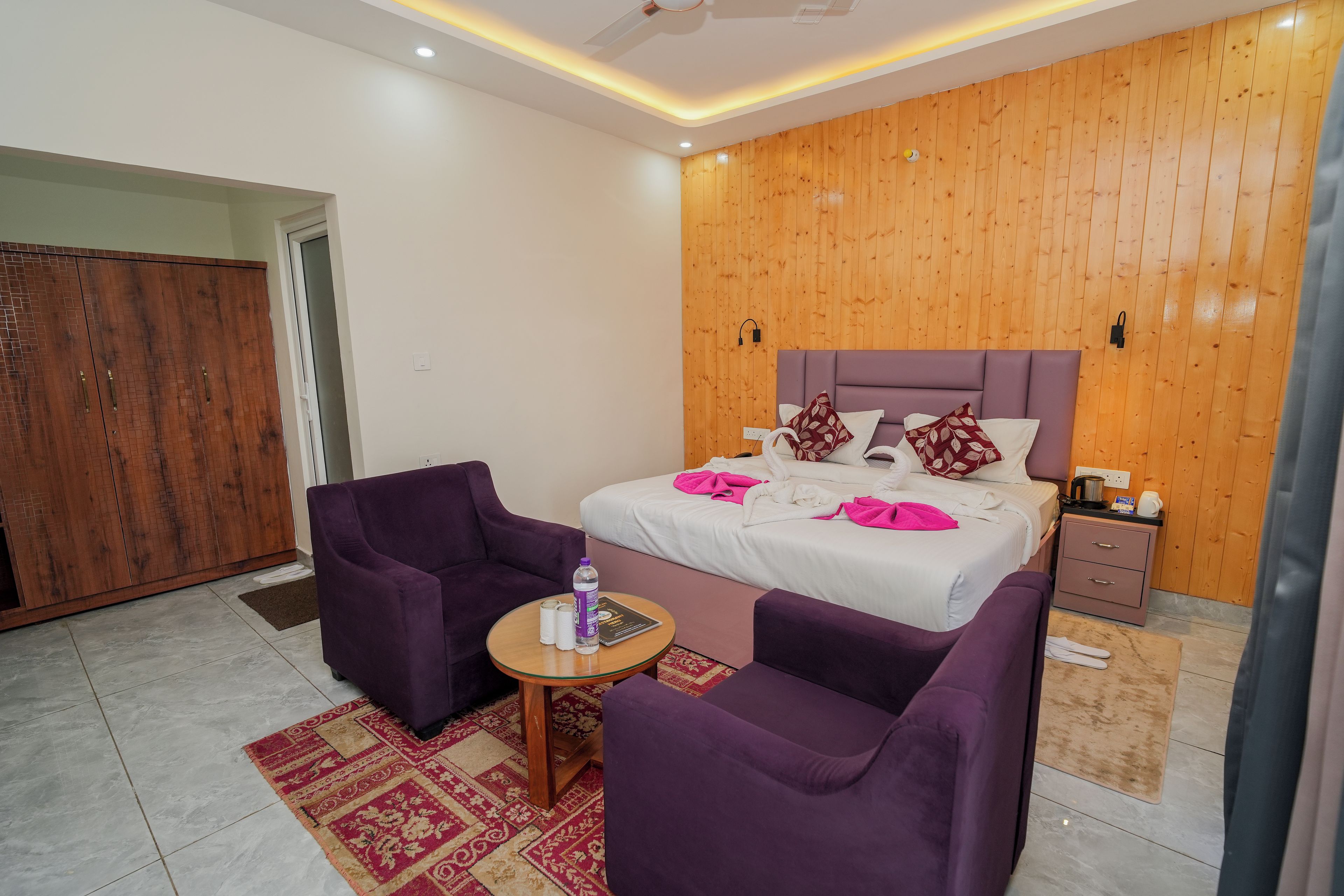 A modern hotel room with a wooden wall, a bed with white bedding, purple armchairs, a small round table with a bottle, and a nightstand with a kettle.