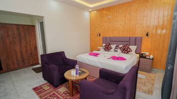 A modern hotel room with a wooden wall, a bed with white bedding, purple armchairs, a small round table with a bottle, and a nightstand with a kettle.