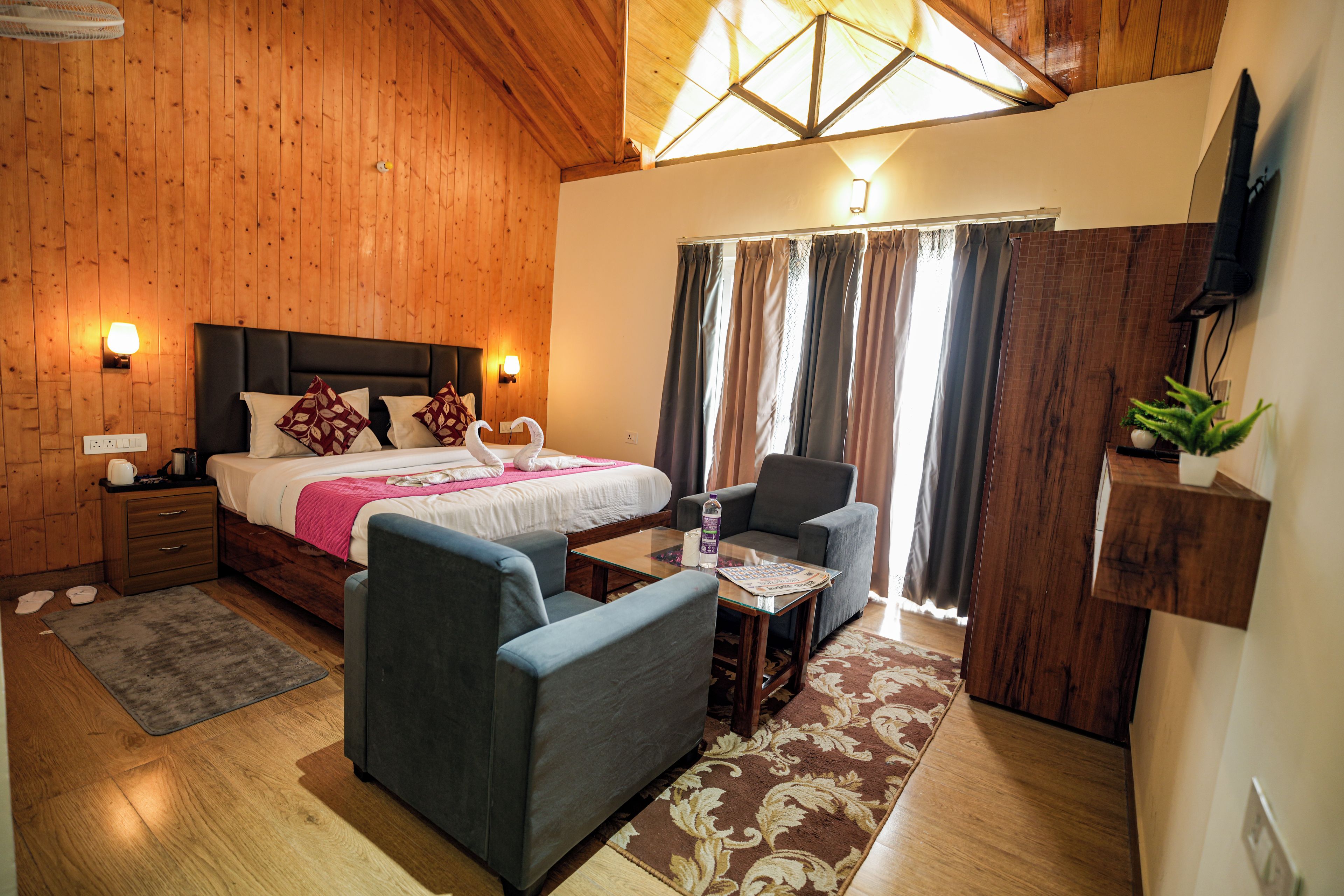 A hotel room with a wooden ceiling, a bed with pillows, a small table with a bottle, a chair, and a TV mounted on the wall.