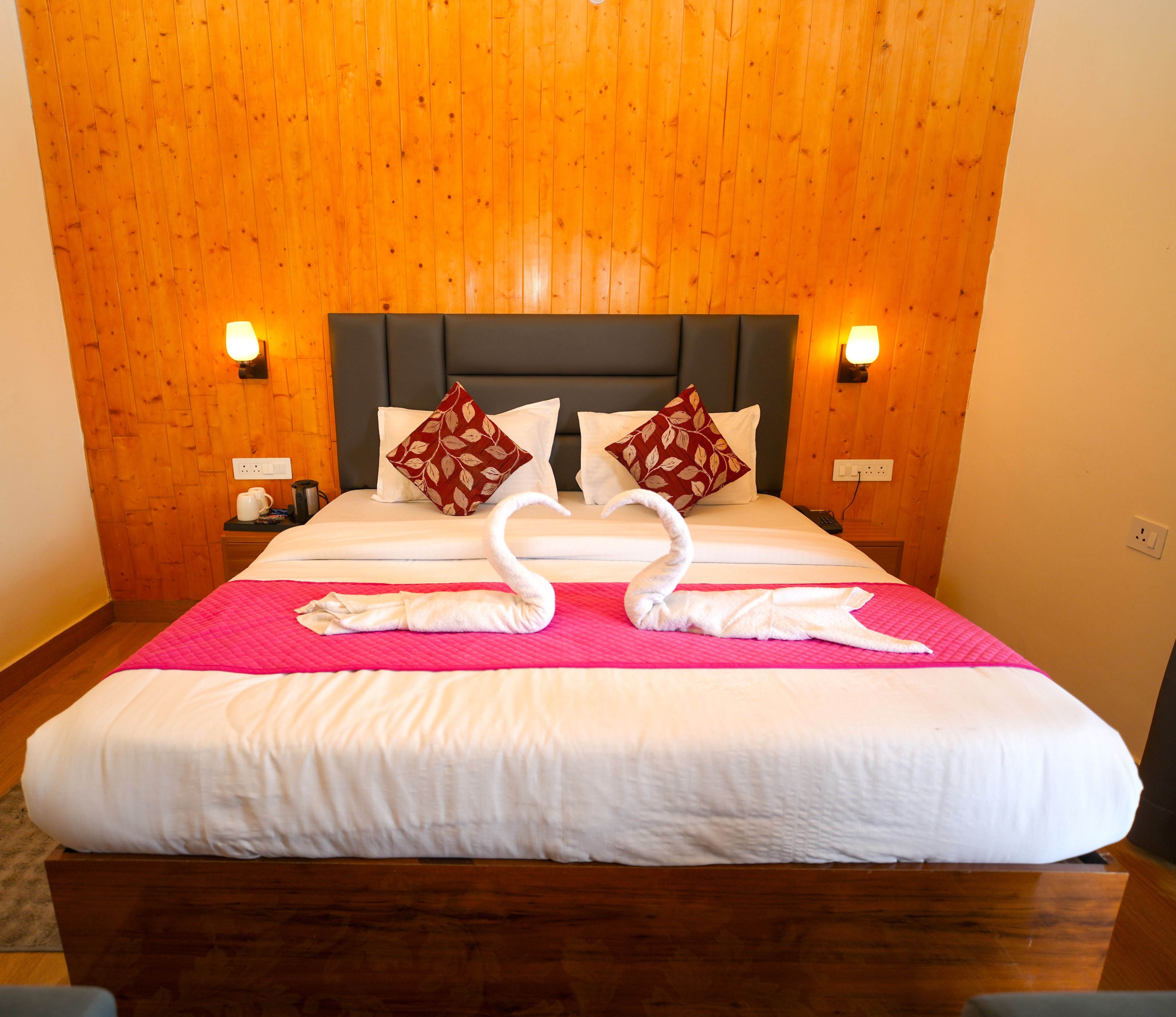 A hotel room with a wooden paneled wall, a bed with white bedding, two decorative pillows, a pink bedspread, and two towels arranged in a heart shape.