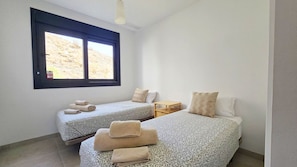 A room with two beds, each with patterned bedding and towels, a small wooden nightstand, and a window with a view of a hillside. - Luxury 2-Bedroom Apartment in El Higuerón (Fuengirola)