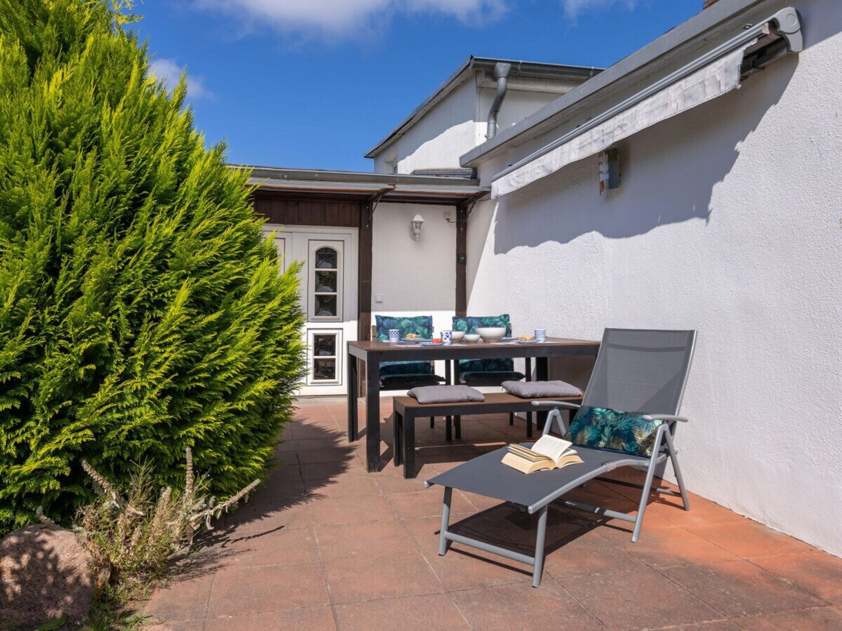 A patio with a table and chairs, a lounge chair with a book, and a large evergreen tree.