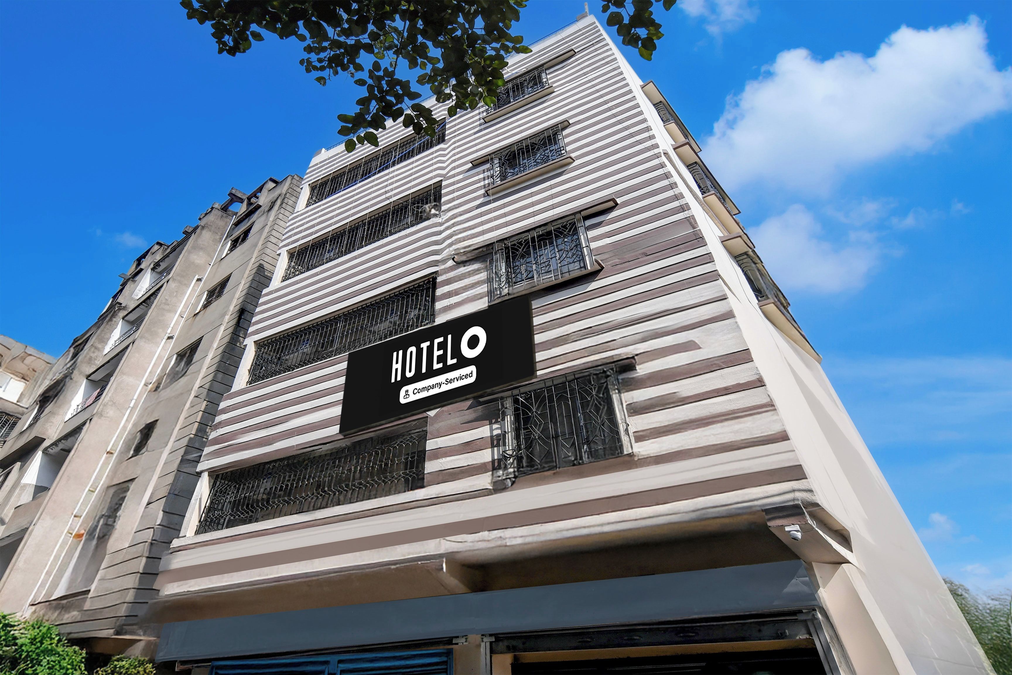 A hotel building with a sign indicating it is company-serviced. The building has multiple floors with barred windows and a modern architectural style.