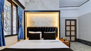 Ein Hotelzimmer mit einem großen Bett, einem mit Beleuchtung versehenen Kopfteil, einem Nachttisch, einer Topfpflanze und Blick durch ein mit Vorhängen drapiertes Fenster.