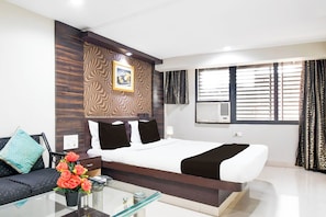 A hotel room with a large bed, a sofa, a glass coffee table with a vase of flowers, a wall-mounted air conditioning unit, and a window with curtains. - Hotel O Raipur formerly Hotel Okasu (Raipur)