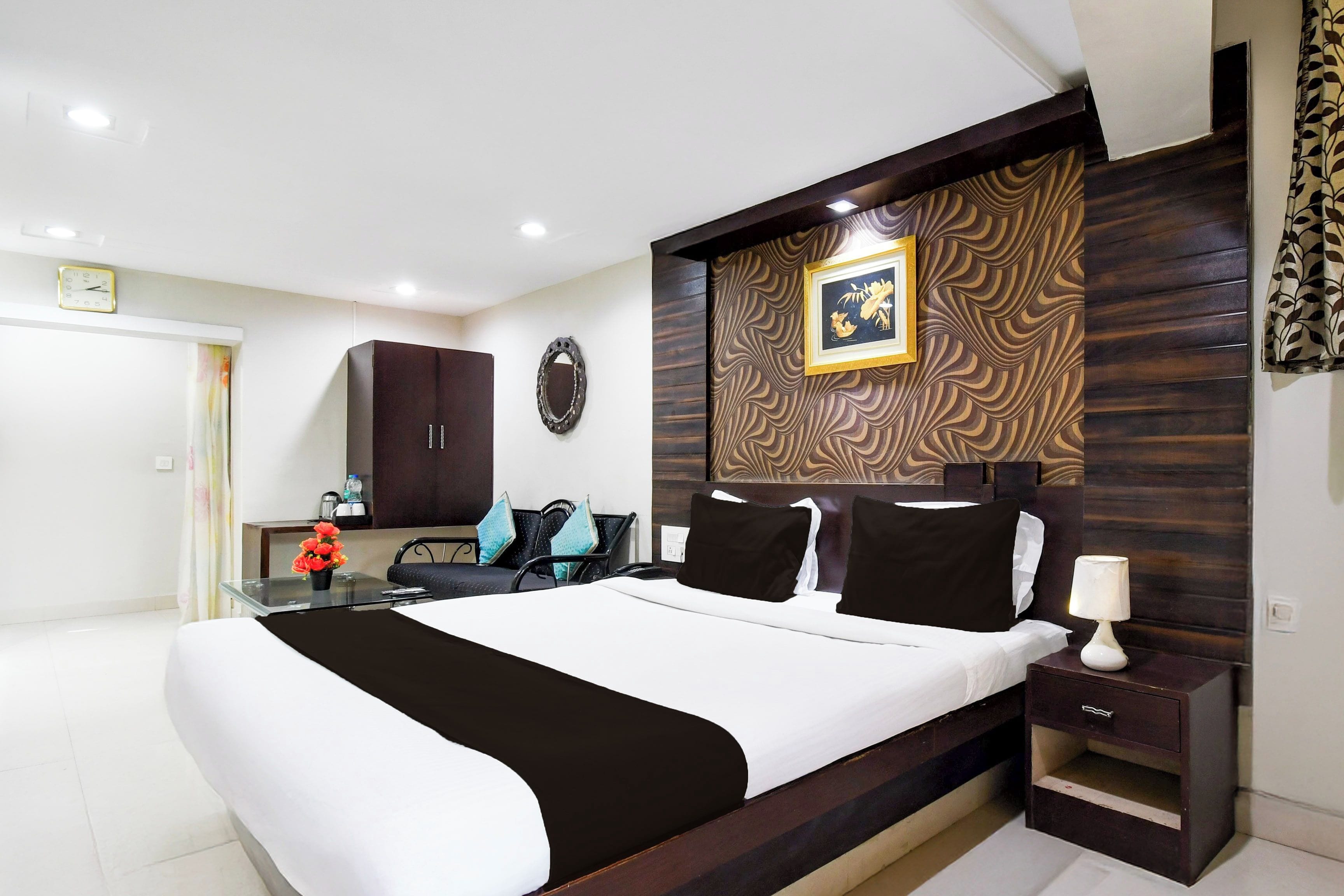 A modern bedroom with a large bed, a bedside table, a desk, a chair, a mirror, and a clock on the wall.