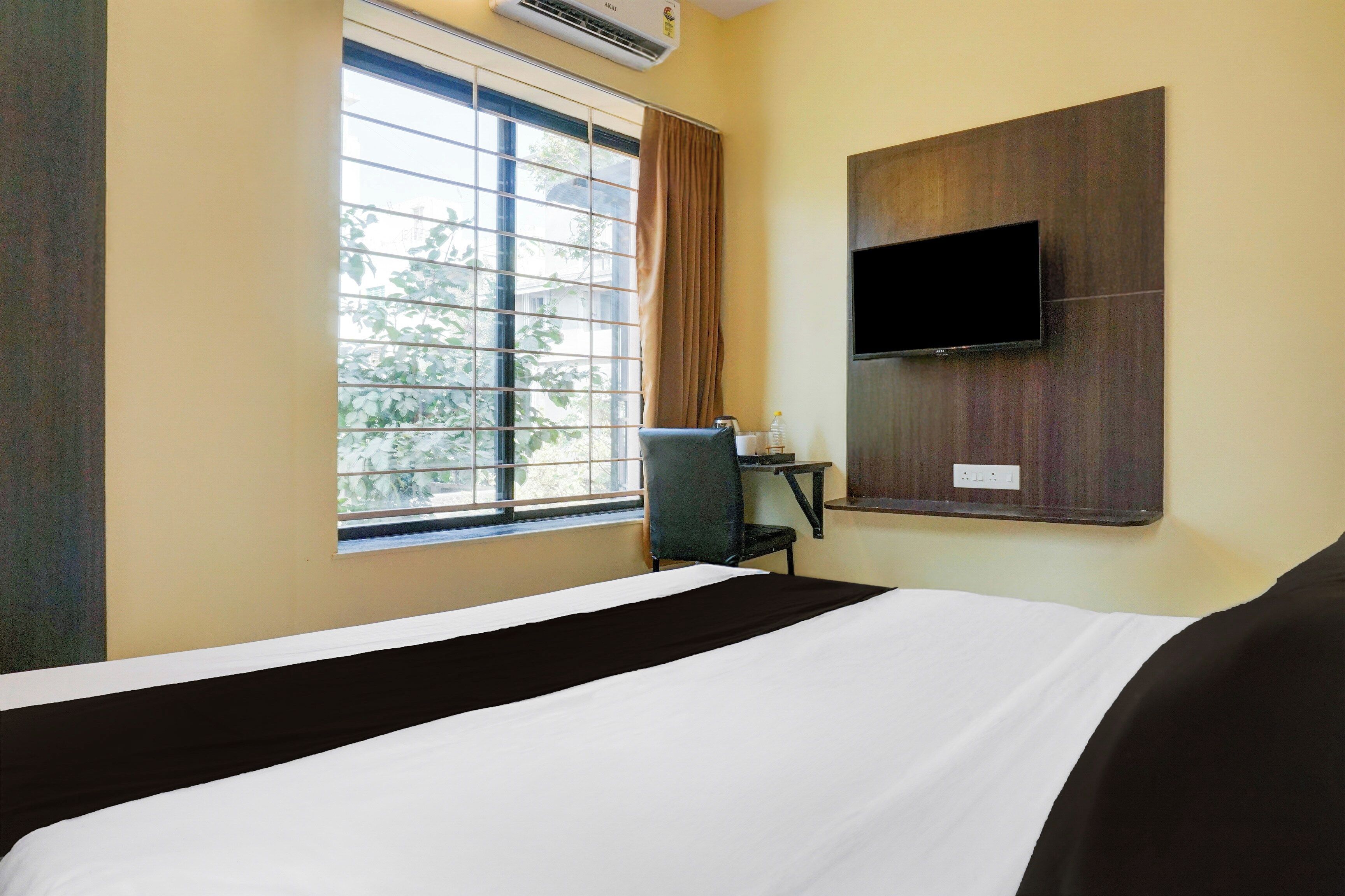 A hotel room with a bed, a desk with a chair, a window with curtains, and a flat-screen TV mounted on the wall.