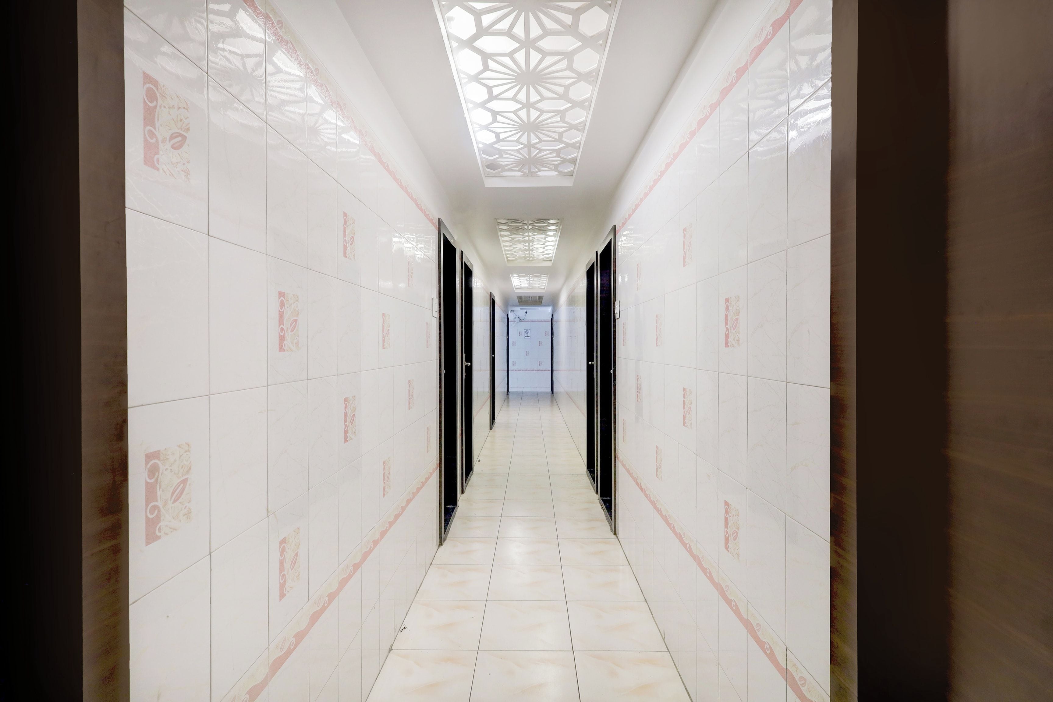 A corridor with white tiled walls and floor, featuring a geometric ceiling design and doors on both sides.