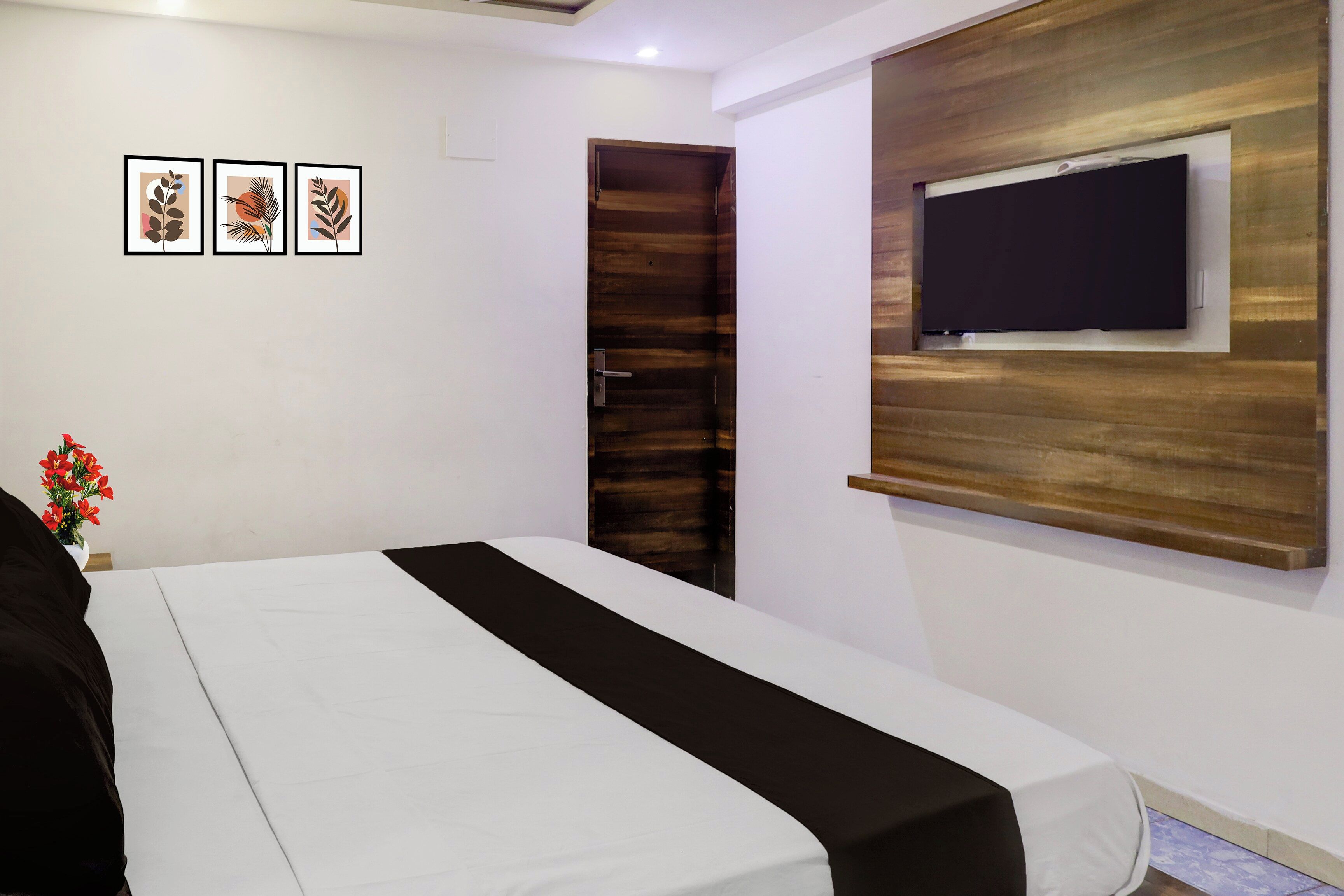 Une chambre d’hôtel avec un lit, une porte en bois, un téléviseur fixé au mur et trois œuvres d’art encadrées accrochées au mur.