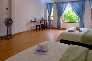 A room with two beds, a desk with chairs, a fan, and a window with curtains.