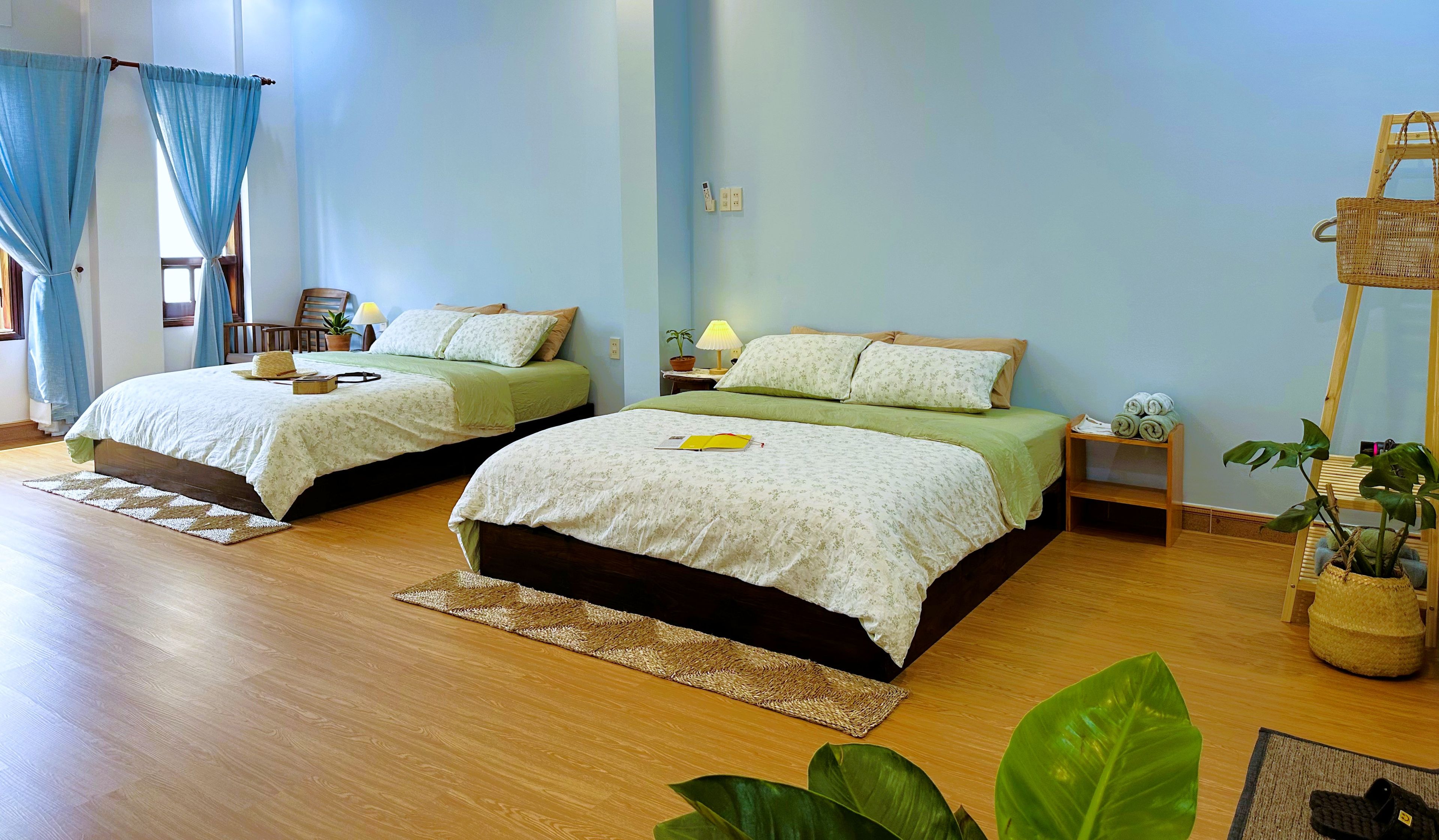 A hotel room with two beds, a wooden floor, a small table with a lamp, and a potted plant.