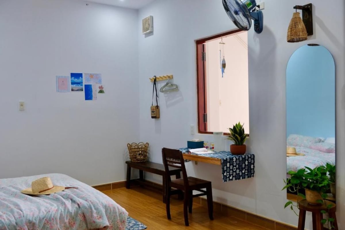 A bedroom with a bed, a desk, a chair, a window, and a wall-mounted fan.