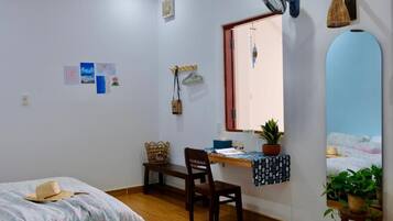 Un dormitorio con cama, escritorio, silla, ventana y ventilador de pared.