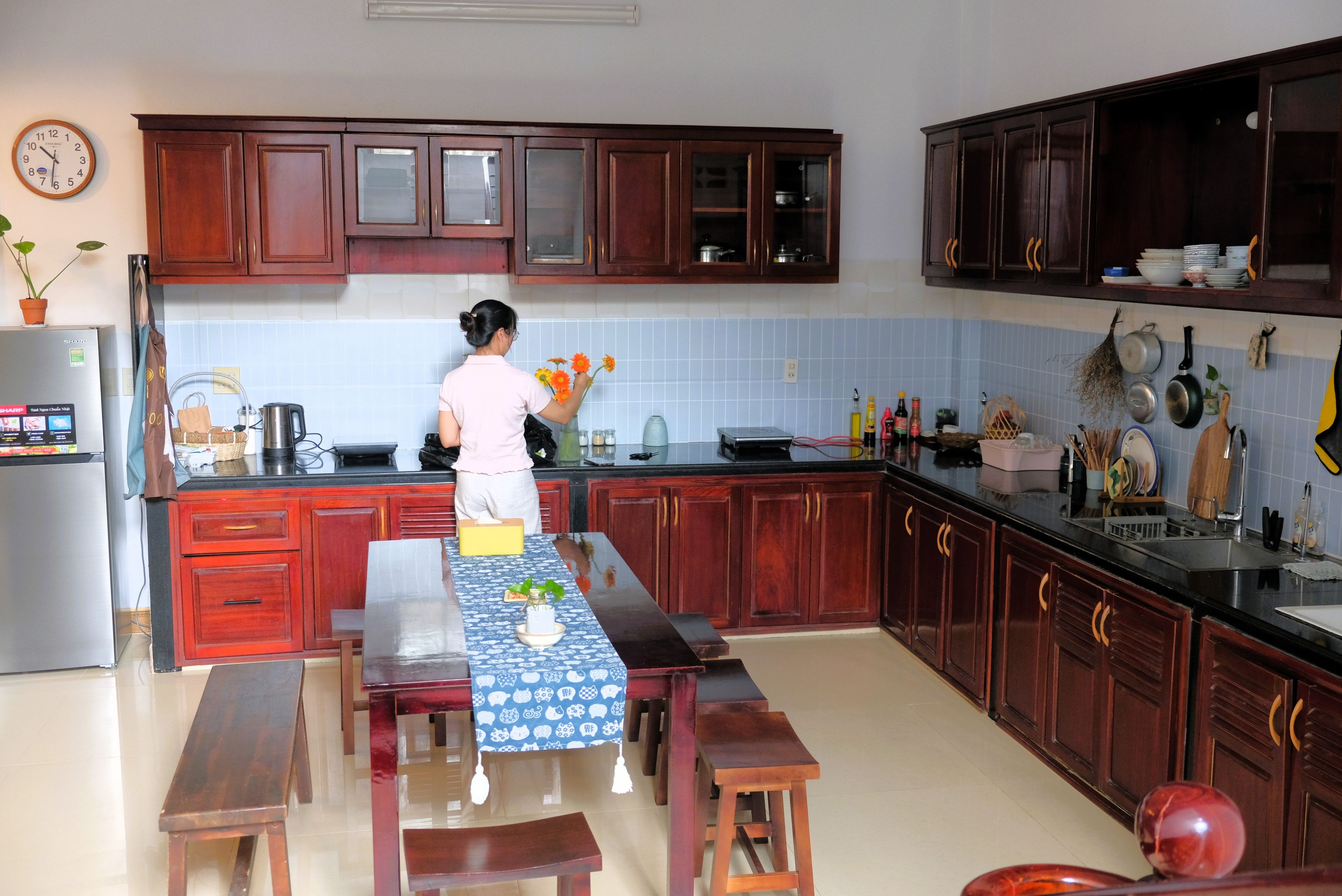 A kitchen with wooden cabinets, a refrigerator, a dining table with benches, and a clock on the wall.