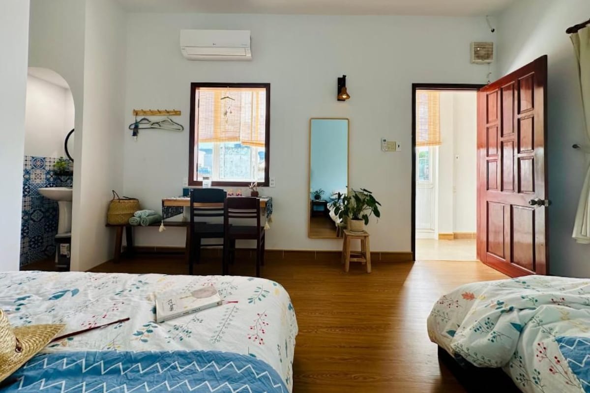 A room with two beds, a desk, a chair, a window with blinds, an air conditioner, and a door.