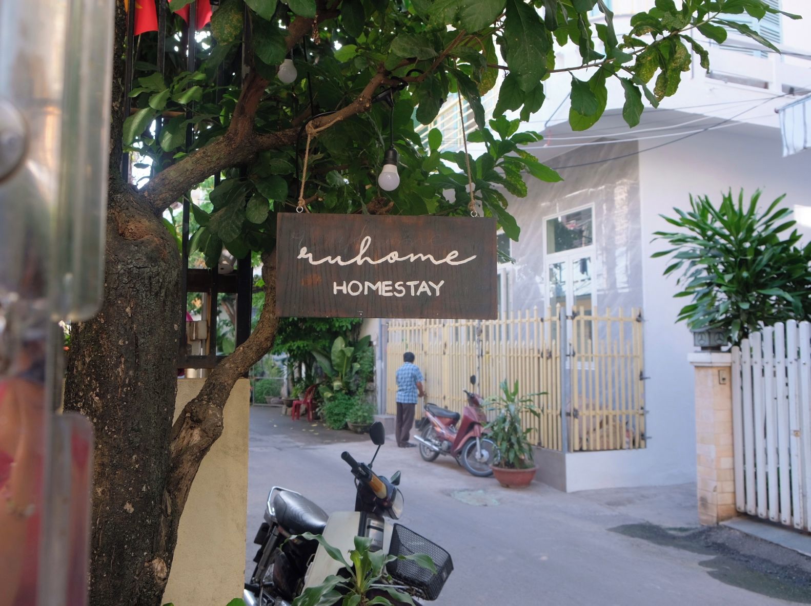 Placa com a inscrição 'home HOMESTAY' pendurada em uma árvore, uma scooter estacionada em frente a um prédio, e uma pessoa caminhando ao fundo.