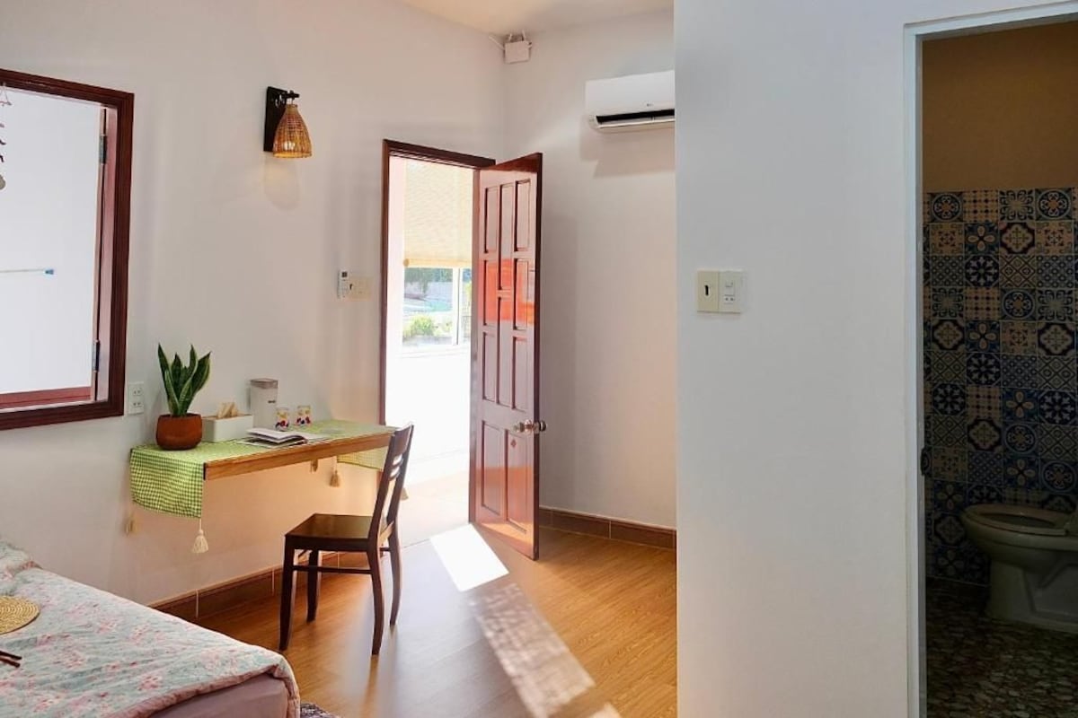 A room with a bed, a desk with a chair, a potted plant, and a door leading to another room.
