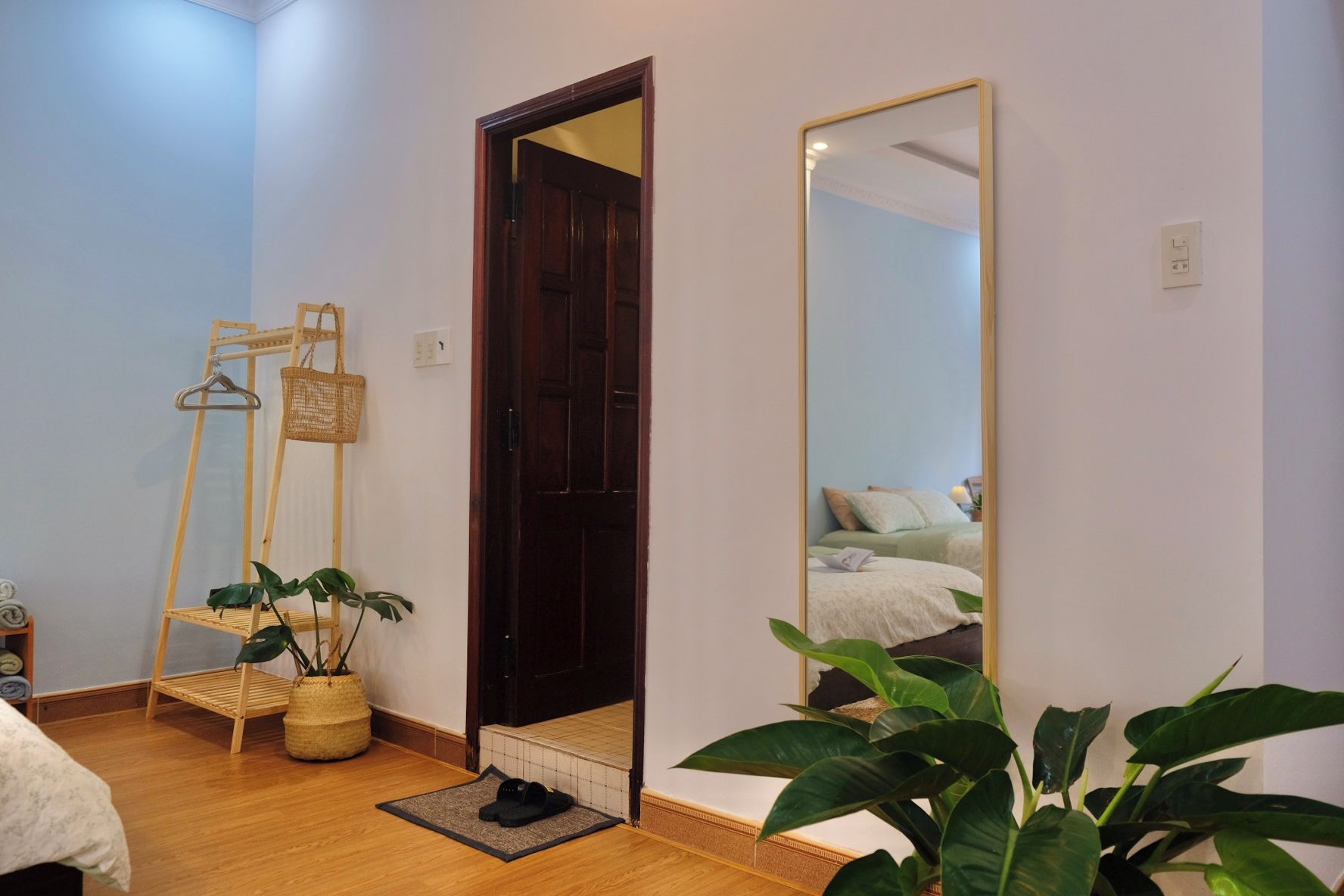 Quarto com porta de madeira, um espelho grande, cama com almofadas, uma planta e uma pequena prateleira com uma cesta.