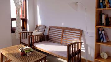 Quarto com sofá de madeira, uma mesinha com uma planta, estante de livros e janela com cortinas.