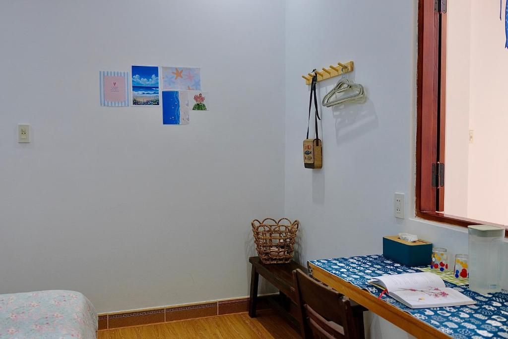A small, tidy room with a single bed, a wooden table with a blue tablecloth, a wicker chair, and a wall-mounted coat rack.