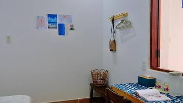 A small, tidy room with a single bed, a wooden table with a blue tablecloth, a wicker chair, and a wall-mounted coat rack.