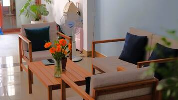 A room with two sofas, a table with a vase of flowers, a fan, and a suitcase against the wall.