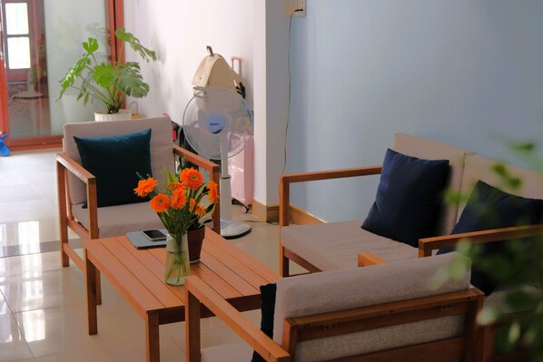A room with two sofas, a table with a vase of flowers, a fan, and a suitcase against the wall.