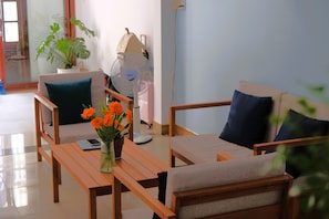 A room with two sofas, a table with a vase of flowers, a fan, and a suitcase against the wall.
