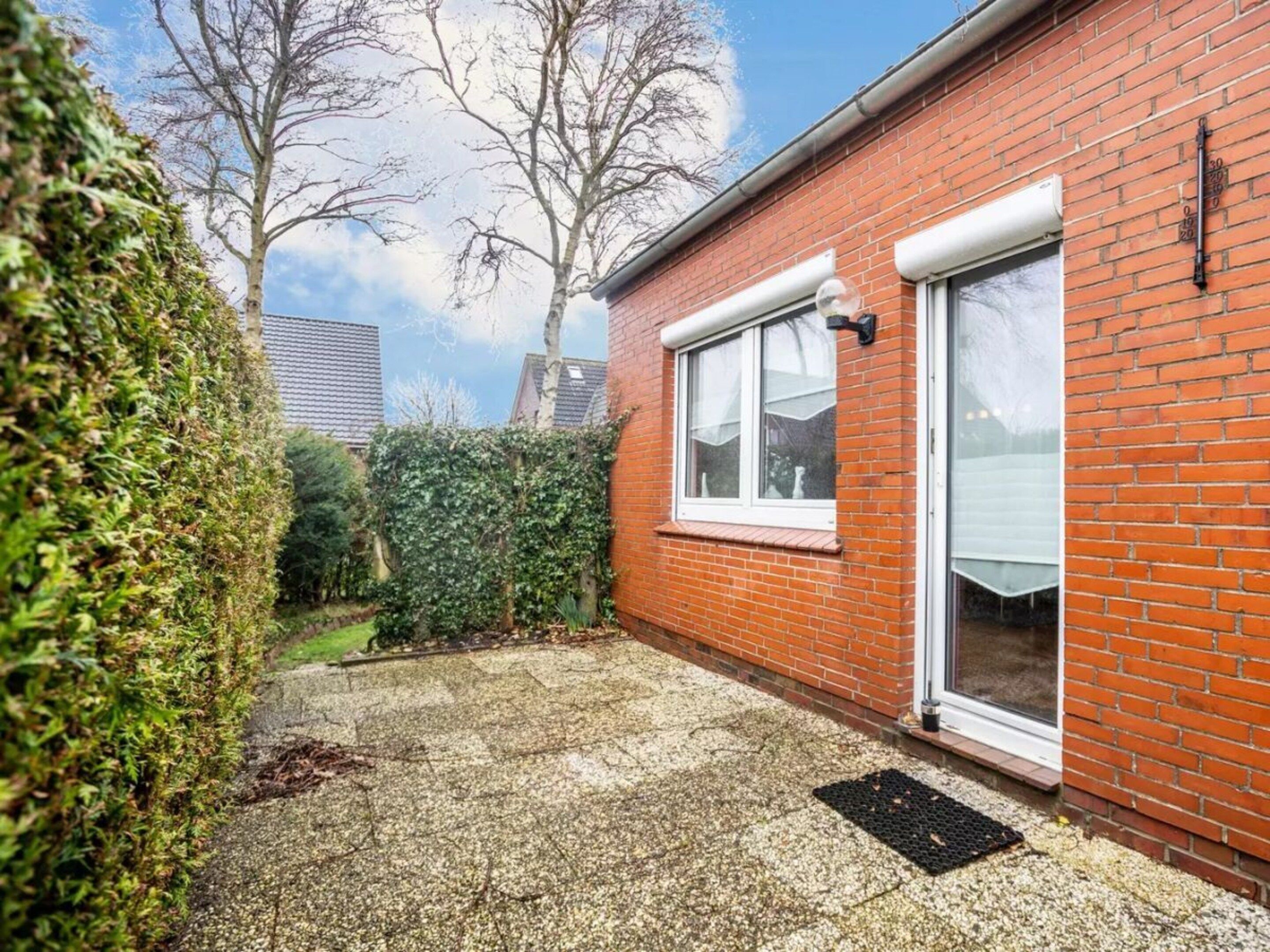 A brick house with a white door, a window, and a small garden area with a hedge and a tree.