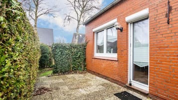 A brick house with a white door, a window, and a small garden area with a hedge and a tree.