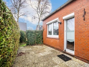 A brick house with a white door, a window, and a small garden area with a hedge and a tree. - Niedl App Helene 2 (Dornum)