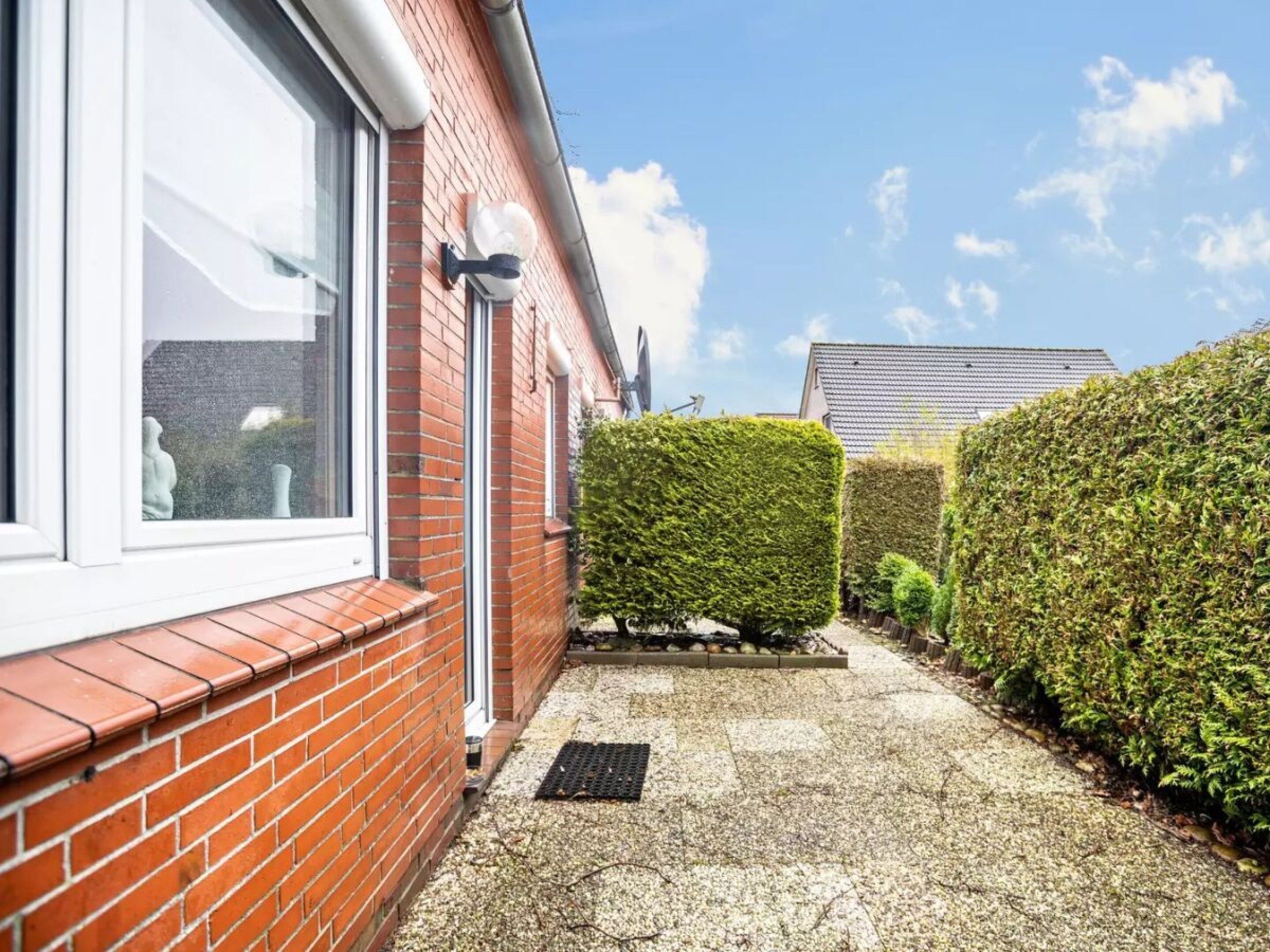 A red brick house with a white window and door, a small garden with a neatly trimmed hedge, and a clear blue sky.