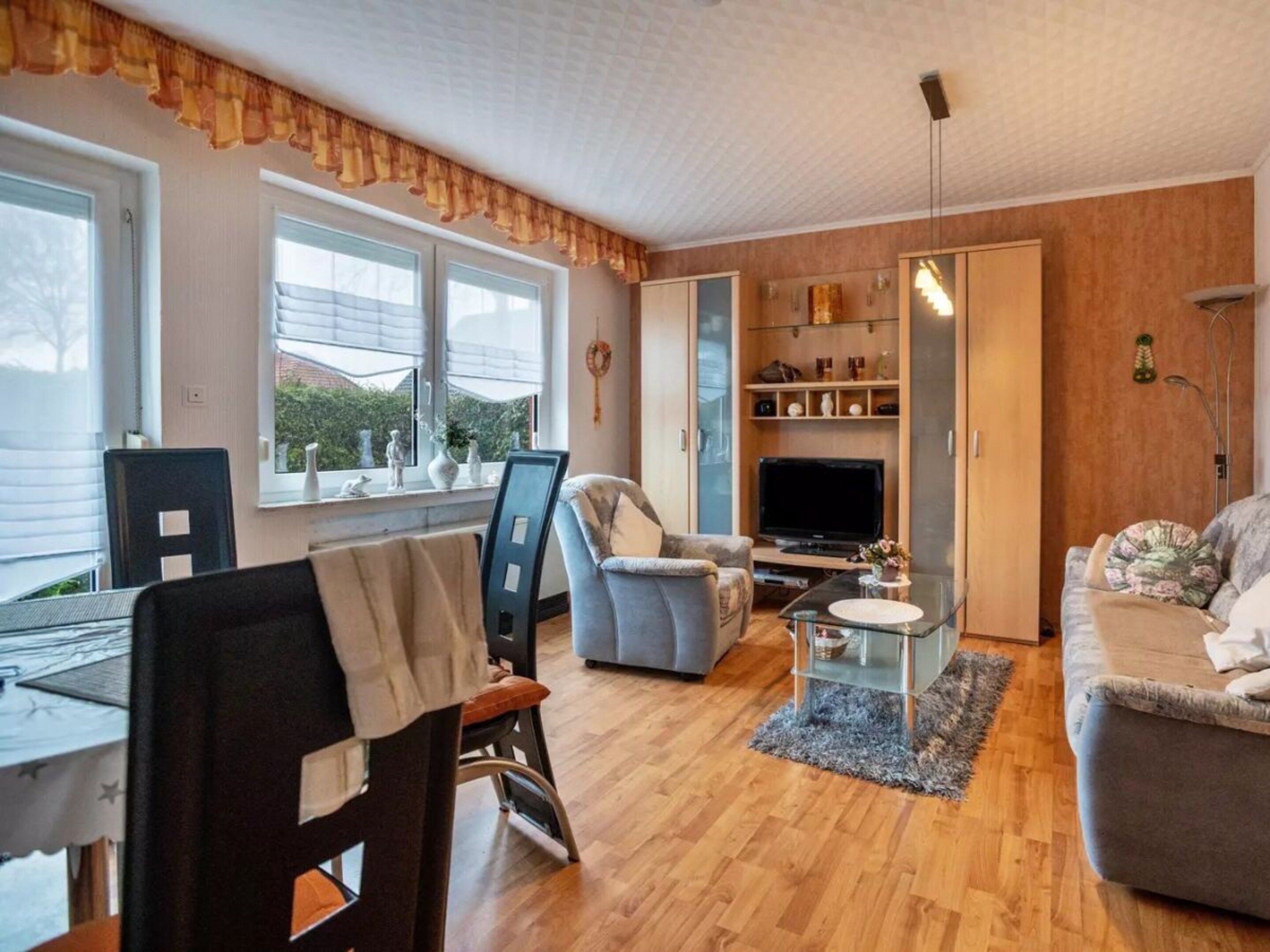 A cozy living room with a sofa, armchair, and a TV stand. There is a dining table with chairs and a window with blinds.