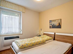 Un dormitorio con cama, ventana con persianas, radiador, un cuadro colgado en la pared y mesita de noche con lámpara.