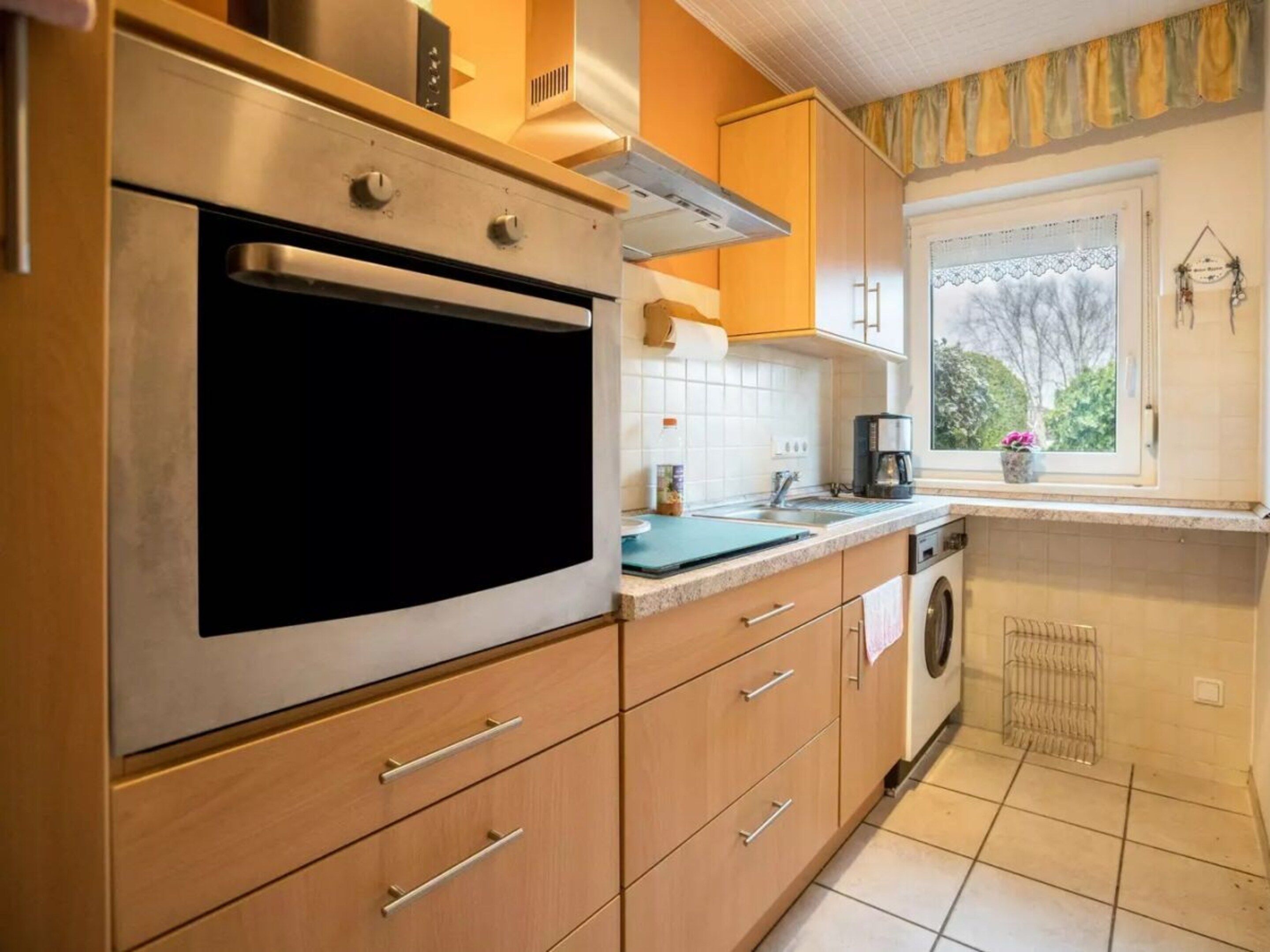 A kitchen with a stainless steel oven, wooden cabinets, a window with a view, and a washing machine.